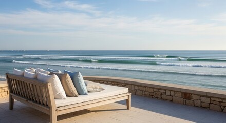 Outdoor lounge with pillows on a stone patio beachfront view of serene waves and distant sailboats beneath a clear sky