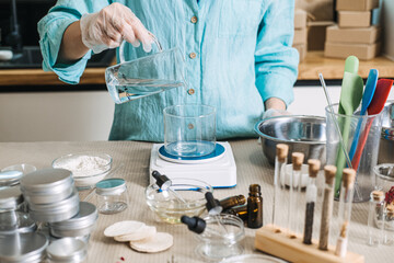 Fototapeta premium Person pours clear liquid from glass pitcher into glass beaker on digital scale. Cosmetic emulsion physics, oil and water mixing, skincare manufacturing science, professional cream base making.