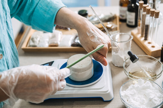 Person applies white cream to back of hand above jar on digital scale surrounded by skincare ingredients. barrier repair cream, precision dosing actives, minimalist formulation, smart scale DIY.