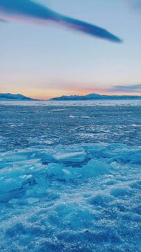 A stunning vertical shot of the "Blue Ice" phenomenon at Sayram Lake, Xinjiang, China. Clear blue ice blocks are surging and crashing onto the shore driven by lake waves. The background features snow-