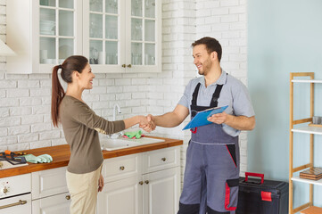 Obraz premium Professional cheerful repairman in uniform shaking hands with happy smiling woman after successful repair standing in kitchen. Man plumber making a deal with client. Plumbing maintenance concept.