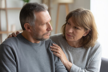 Obraz premium Mature couple sitting together at home, woman gently comforting thoughtful man with hand on shoulder, emotional support, relationship care, empathy and warm indoor atmosphere.