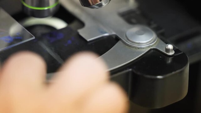 Close up laboratory video footage showing controlled movement of microscope stage while observing specimen on glass slide. Precise mechanical adjustment used in medical research diagnostics histology
