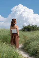 a woman stands on the path, facing away with her back. she is standing in tall grass and looks up at the sky where there is one large cloud. she wears a brown skirt and a white top.