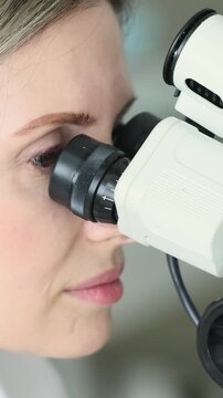 Close up of mature female scientist with fair skin and gray hair conducting research using black laboratory microscope. Woman carefully adjusts eyepieces while examining specimen during scientific