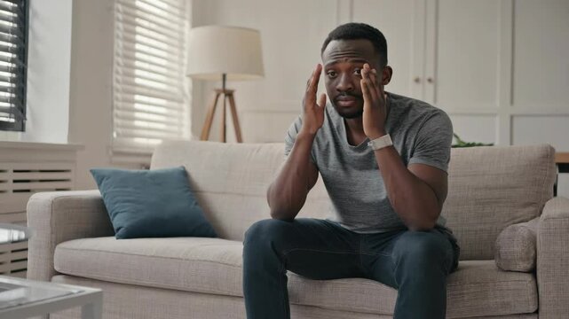 A man in emotional pain sits alone on the couch covering his face with both hands.