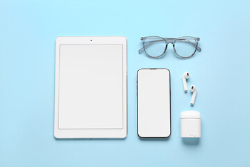 Modern tablet computer, mobile phone, earphones and eyeglasses on blue background, closeup