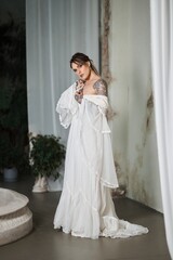 Tattooed woman in flowing white dress posing elegantly in a vintage interior setting