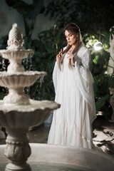 Elegant woman in a white flowing gown stands by a tiered stone fountain in a lush garden setting