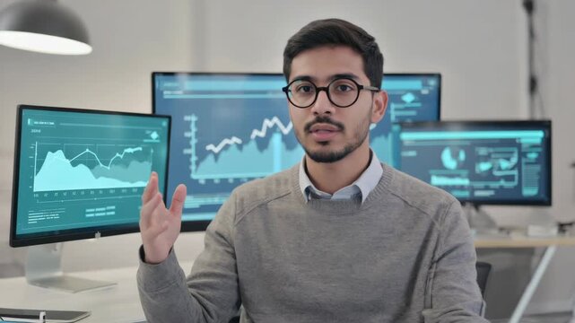 A man confidently gestures towards multiple monitors displaying charts and graphs, indicative of a financial or business environment.