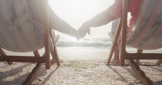 Back view of hispanic senior couple relaxing on sunbed on beach at sunset, holding hands