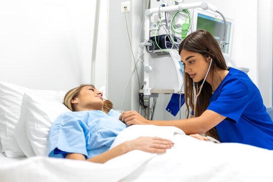 A young female doctor or nurse in blue scrubs uses a stethoscope to examine a patient lying in a hospital bed. Professional medical consultation, diagnostic checkup, and attentive clinical care.