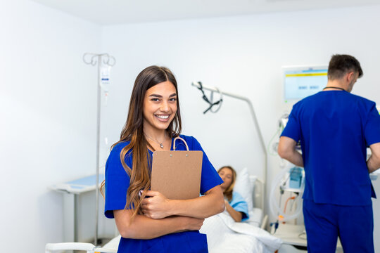 Smiling female nurse with clipboard in hospital ward. Professional medical staff, Healthcare And Medicine concept. Portrait of happy young doctor in blue scrubs with patient in background.