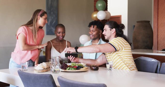 At party, four friends laughing and sharing smartphone at dining table with snacks and drinks, ballo
