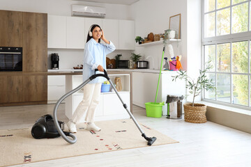 Fototapeta premium Young woman listening to music while vacuuming carpet in kitchen