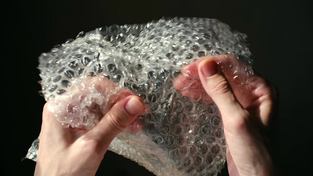 Hands popping bubble wrap on dark background, satisfying asmr, anti stress activity. Close up showing fingers bursting plastic packaging, creating relaxing sound. Popping bubble wrap for stress relief