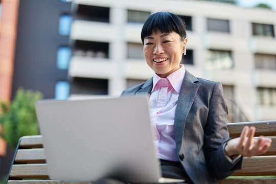 Portrait of a young asian japanese businesswoman woman using a laptop computer having a video call conference and sitting on a bench in a coty park, surrounded by modern corporate office buildings