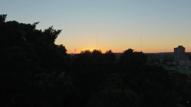 Drone ascends above dense treetops to reveal Puerto Iguaz&uacute; cityscape at sunset, with clear sky and golden horizon