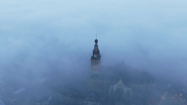 Aerial view of the Stevenskerk standing above Nijmegen during a foggy sunrise, with soft mist drifting through the narrow streets and warm light coloring the sky. The church tower appears from the haz