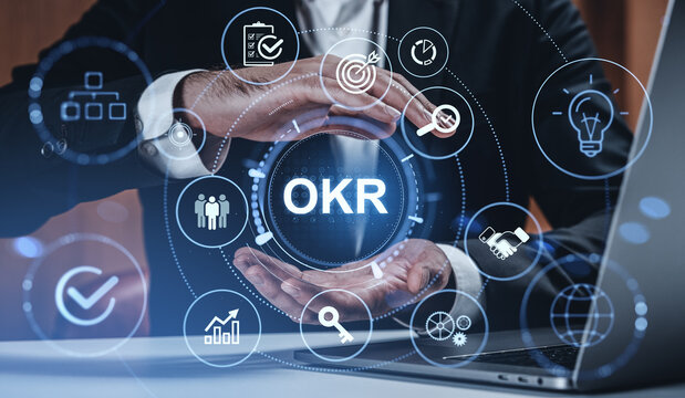 Businessman holding virtual OKR icon surrounded by digital symbols of goals, teamwork, and analytics on creative tech background in office setting.
