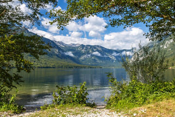 Clear Bohinj lake (Bohinjsko jezero) reflects mountains under a blue sky in Slovenia. Trees frame the scene along the water's edge. Nature shows its beauty.