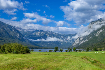 The scene shows Bohinj Lake (Bohinjsko jezero) in distance surrounded by  mountains under a blue sky with clouds. Nature inviting for hiking and exploration.