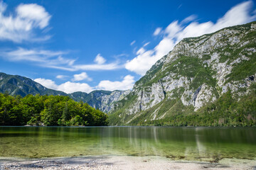 Bohinj lake (Bohinjsko jezero) shows clear water surrounded by green trees and mountains under a blue sky with clouds. Nature thrives in this area of Slovenia.