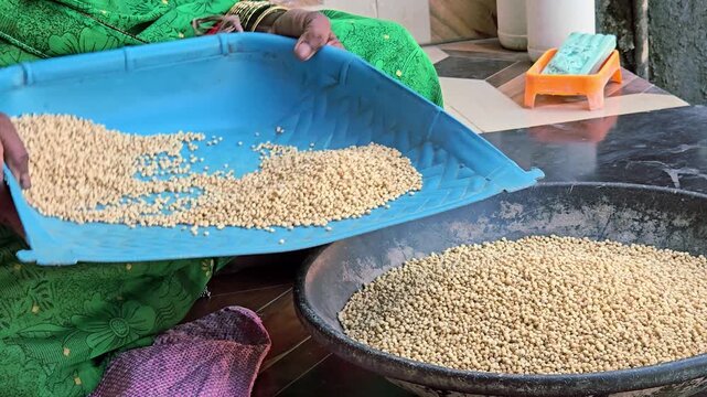 Rural India: an Indian woman housemaker performs the skilled, traditional dance of winnowing jowar, separating chaff from grain using a blue tray, an essential domestic act of care to prepare a meal.
