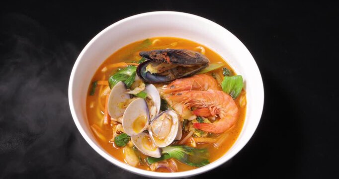 Against a black background, a close-up photo showcases a steaming bowl of Korean spicy seafood soup noodles filled with fresh shrimp, mussels, clams, and vegetables.