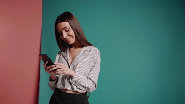 Young stylish woman with long brown hair looking down and smiling while actively engaging with a modern smartphone device indoors against vibrant studio backdrops