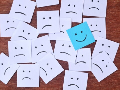 Blue smiling face adhesive note among many white sad faces on wooden background. Positivity Concept for optimism, mental health awareness and staying positive
