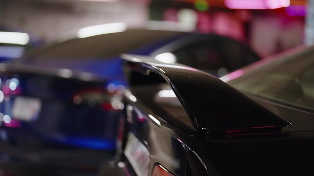 Shiny black spoiler of a modern sports car parked inside an underground garage, with colorful blue and pink neon lights reflecting off the glossy paint and blurred vehicles in the background