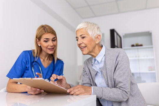 Smiling female doctor holding a clipboard while a happy senior patient signs a medical consent form or insurance document during a clinical visit.