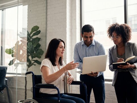 Diverse business team with woman in wheelchair discussing project on laptop in modern office