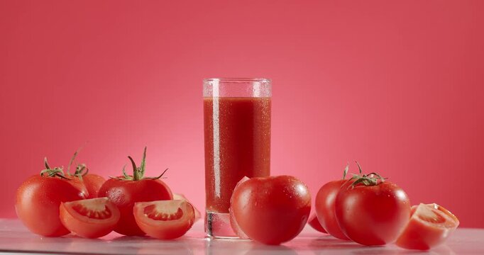 Red ripe tomatoes and a glass full of tomato juice rotating on a glass surface against a red background, showcasing a healthy and refreshing vegetarian and vegan beverage in slow motion