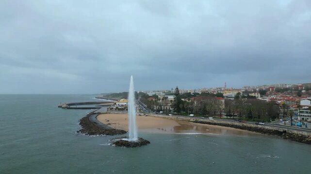 Circular aerial view of the fountain in the sea with the waterfront and the town of Pa&ccedil;o de Arcos in the background on a cloudy day.Portugal.