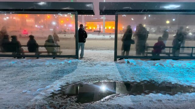 The video opens with a panoramic view of an illuminated bus stop at dusk: the streetlights cast a yellow glow on the snow covering the sidewalk and the roof of the pavilion. Tired passengers in warm 
