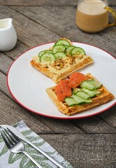 Oatmeal waffles with savory toppings: hummus, fresh cucumber, and salted salmon on a white plate against a wooden background.