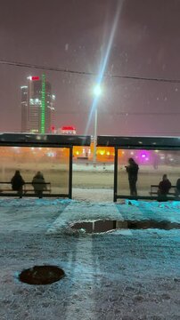 The video opens with a panoramic view of an illuminated bus stop at dusk: the streetlights cast a yellow glow on the snow covering the sidewalk and the roof of the pavilion. Tired passengers in warm 