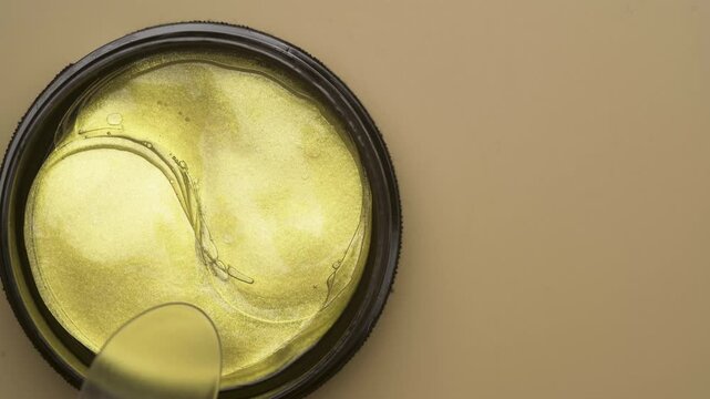Takes gold eye patches with plastic spoon from jar on beige background, top view. Cosmetic product