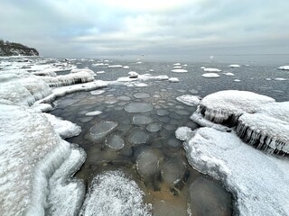 A winter shoreline stretches toward a calm horizon, the foreground is filled with frozen water patterned by circular ice plates locked together like a mosaic and snow-covered rocks