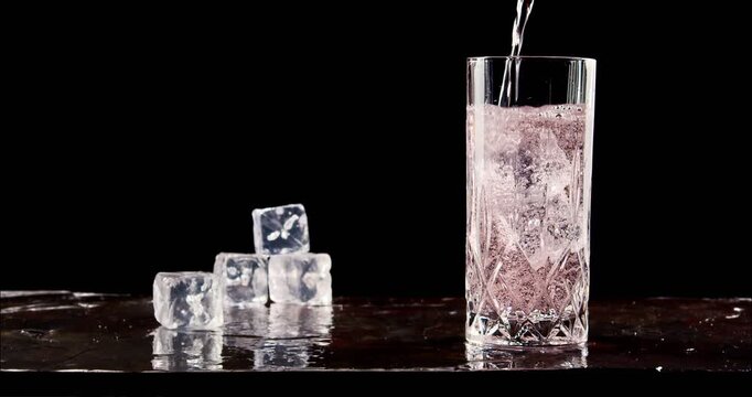 Pouring cold drink to glass with ice. Slow motion, copy space, black background. Hard seltzer, sparkling water, gin tonic, soft drink.