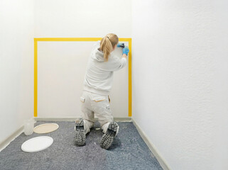 Handwerkerin bei Malerarbeiten in einem Innenraum. Die Frau kniet auf dem Boden und klebt mit Kreppband einen rechteckigen Bereich an der Wand ab, um saubere Farbkanten zu erzielen
