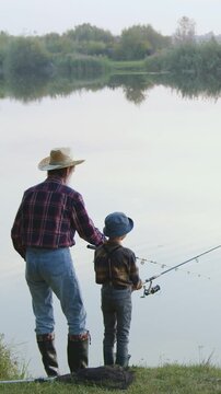 Portrait of attractive concentrated mature bearded grandfather in straw hat and small interested grandchild which standing together on the pond's bank and throwing rods to catch the fish on hooks with