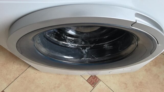 The washing machine is cleaning clothes with soap and water in a home laundry room. Bubbles form in the glass door as the cycle runs. The setting shows a typical weekday afternoon.