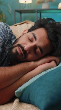 Tired Indian man lying down in bed taking a rest. Carefree young Hindu boy napping, falling asleep on comfortable sofa with pillows at home apartment room. Closed his eyes enjoy daytime nap alone