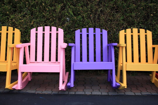 Four colorful Adirondack rocking chairs in pastel pink, purple, and yellow lined up against a green hedge.