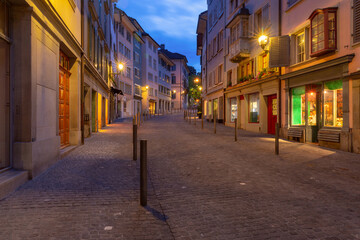 Obraz premium Early morning view of an empty cobblestone street in Zurich old town, lined with traditional buildings and softly glowing streetlights