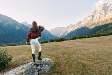 Fototapeta premium Woman standing on a rock in a grassy meadow overlooking mountains and a valley at sunset. Traveler in brown jacket and boots surveys scenic alpine landscape and distant peaks.