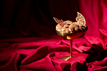 Obraz premium A gourmet tiramisu dessert is elegantly served in a gold coupe glass, garnished with chocolate filigree and coffee beans, set against a luxurious draped red velvet backdrop.